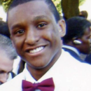 A headshot of a smiling person wearing a white shirt and a red bow tie, with a blurred outdoor background.