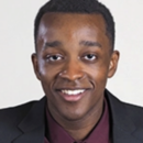 A headshot of a person wearing a dark suit and a maroon shirt, smiling, with a light neutral background.