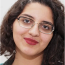 A person with curly hair and glasses, wearing red lipstick, smiling slightly against a plain background. This is a headshot.