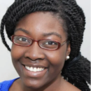 Headshot of a person with glasses and braided hair, smiling. They are wearing a blue top.