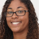 A headshot of a smiling person with curly hair and glasses against a plain background.