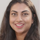 Headshot of a person with long dark hair, smiling broadly. They have prominent eyebrows and are wearing earrings, with a light background.