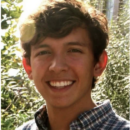 A headshot of a person smiling, with short hair, wearing a plaid shirt, and standing outdoors with greenery in the background.