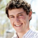 Headshot of a person with curly hair, smiling, wearing a striped shirt, outdoors with blurred greenery in the background.