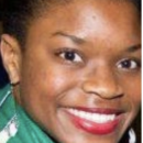 Headshot of a person smiling, wearing a green garment with short, braided hair. The background is blurred.