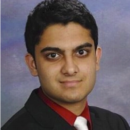 A headshot of a person wearing a dark suit, red shirt, and white tie against a blue-gray background.