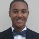 Headshot of a person smiling, wearing a dark suit jacket, white shirt, and a polka-dotted bow tie against a plain background.