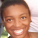 Headshot of a person smiling with hair pulled back, wearing earrings. Background is blurred with outdoor elements.