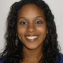 Headshot of a person with long, curly hair, smiling, and wearing earrings. They have a neutral background.