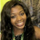 A headshot of a person with long wavy hair, smiling, wearing a dark top and a green lanyard. The background is a mix of green and brown hues.