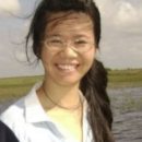 A person with long hair and glasses is smiling outdoors near water and grassy fields. It is a sunny day with a few clouds in the sky.