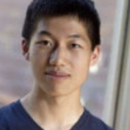 Headshot of a person with short dark hair, wearing a dark V-neck shirt, and standing in front of a blurred, neutral-colored background.