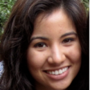 A headshot of a person with long, dark hair and a smile, set against a backdrop of greenery.