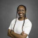 A person with glasses and braided hair, wearing a white knit top, smiling with arms crossed against a gray background.