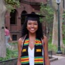 A person wearing a graduation cap and a colorful stole, smiling outdoors with trees and a brick building in the background.