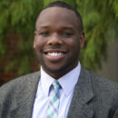 Headshot of a smiling person wearing a suit, tie, and shirt with a blurred greenery background.