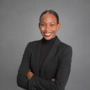 Headshot of a person wearing a black suit and turtleneck, smiling with arms crossed against a gray background.