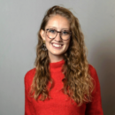 A person with long curly hair wearing glasses and a red sweater, smiling against a plain backdrop.