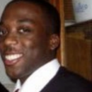 This is a headshot of a person wearing a suit and tie, smiling, with a blurred background.