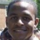 A headshot of a person smiling, outdoors, with trees and a building in the background.