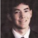 Headshot of a person wearing a suit and tie, smiling at the camera, with a softly blurred dark background.